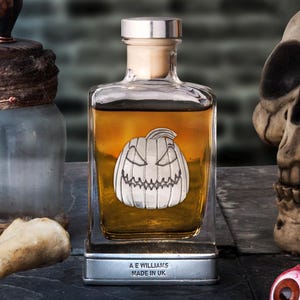 Op de afbeelding: Een Halloween-display met een vierkante glazen fles met amberkleurige vloeistof en een zilveren pompoenontwerp. Andere items zijn antieke glazen flessen, botten, een schedel en een rood oog.