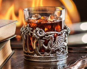 May include: A glass of amber liquid with ice cubes in a silver dragon-shaped holder, sitting on a wooden table with two books stacked next to it. A fireplace with flames is visible in the background.