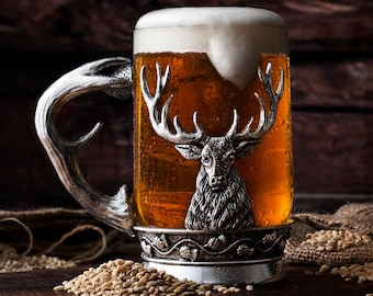 May include: A silver beer stein with a deer head design, filled with a golden beer topped with a white foam head. The stein is sitting on a wooden surface with scattered grains of barley.