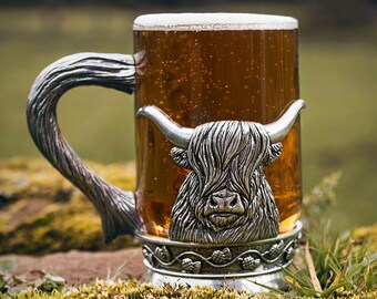 May include: A silver Highland cow mug with a handle, filled with a golden brown liquid. The mug is sitting on a bed of green moss.