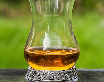 May include: A clear glass whisky tumbler with a gold-colored base, featuring a decorative design of Scottish flags and thistles. The tumbler is filled with amber-colored liquid and is sitting on a dark surface.