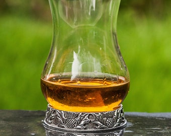 May include: A clear glass whiskey tasting glass filled with amber liquid, resting on a decorative silver base. The base features a thistle design. The glass sits on a wet, dark surface with a blurred green background.