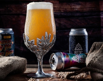 May include: A glass of beer in a pewter goblet with a leafy design, surrounded by hessian sacks, cans of beer, and barley grains on a wooden surface.