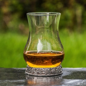 Könnte beinhalten: Ein klarer Whisky-Tumbler aus Glas mit einem silbernen Sockel, der mit einem detaillierten Design verziert ist. Der Tumbler ist mit bernsteinfarbener Flüssigkeit gefüllt und steht auf einer dunkelgrauen Oberfläche.