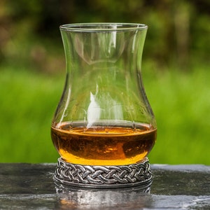 May include: A clear glass whisky tumbler with a silver Celtic knot design base, filled with amber-colored whisky, sitting on a dark gray surface.