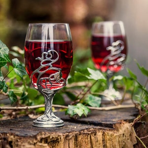 May include: Two wine glasses with silver, ornate, vine-like stems filled with red wine. The glasses are sitting on a wooden stump with green leaves in the background.