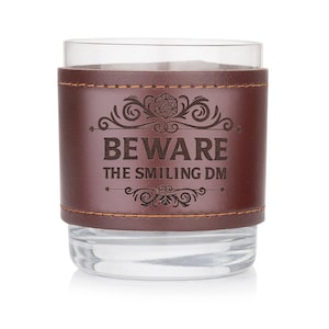 以下が含まれることがあります： 「BEWARE THE SMILING DM」という文字と20面ダイスのグラフィックが刻まれた、茶色の革で覆われたガラス製のタンブラーです。