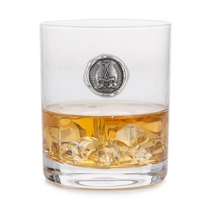 May include: A clear glass tumbler with a silver monogrammed seal on the side. The glass is filled with amber liquid and ice cubes.