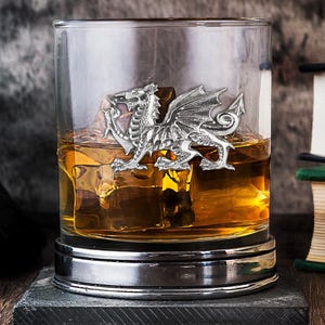 May include: A tumbler glass with a silver dragon emblem, containing amber liquid and ice cubes. The glass rests on a dark coaster, with books in the background. The dragon is intricately detailed.