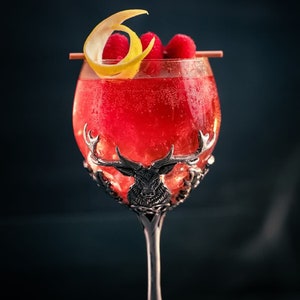 Op de afbeelding: Een rode cocktail met frambozen en een citroenschil in een zilveren kelk met een hertenontwerp.