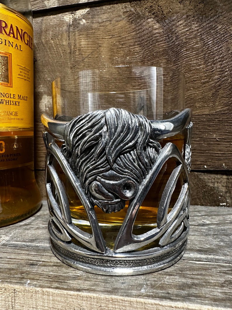 May include: A pewter Highland cow head whiskey glass holder. The holder is in the shape of a Celtic knot with a Highland cow head in the center. The glass holder is designed to hold a standard-sized whiskey glass.