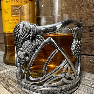 May include: A silver pewter glass holder featuring a Celtic knot design, a bull's head, and a thistle. The holder is designed to hold a glass of whiskey or other spirits.