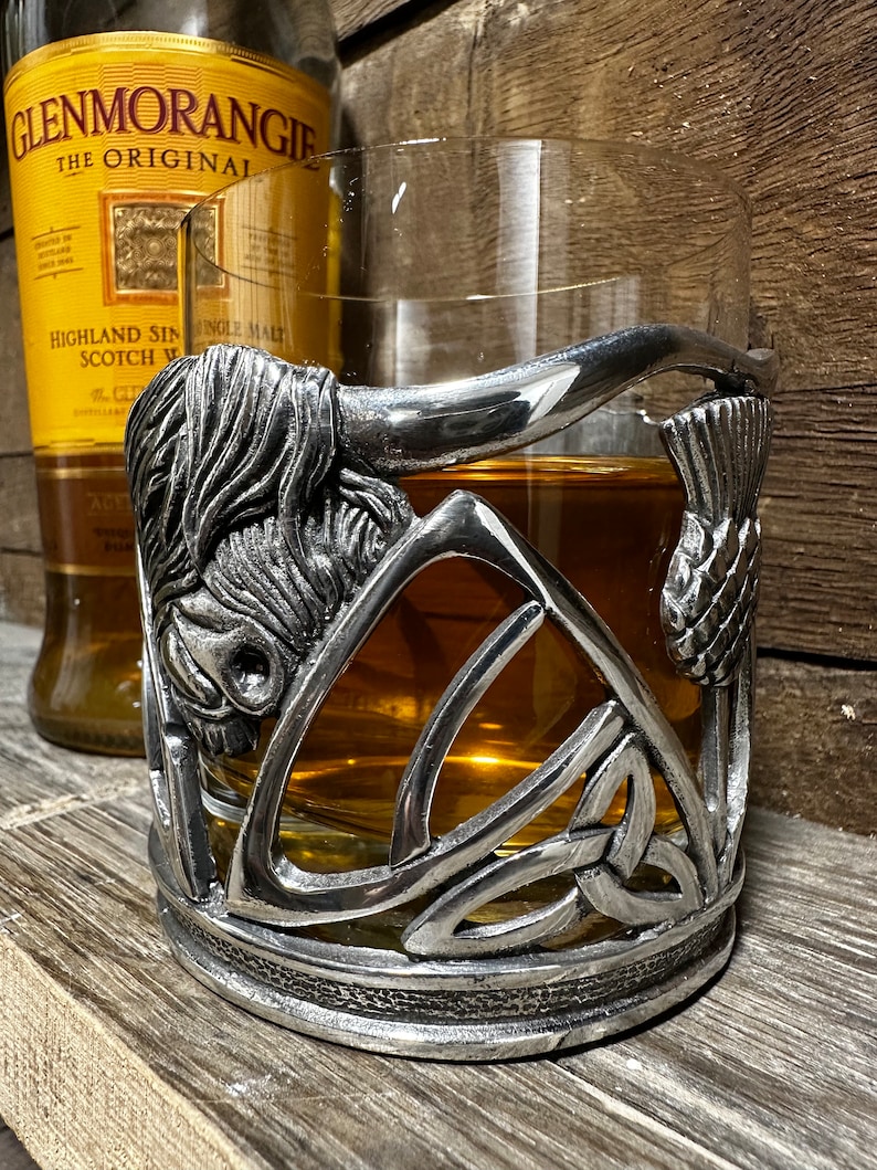 May include: A silver pewter glass holder featuring a Celtic knot design, a bull's head, and a thistle. The holder is designed to hold a glass of whiskey or other spirits.
