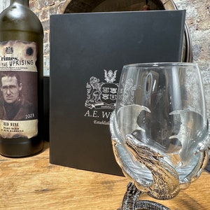 May include: A black box with a silver crest and the text "A.E.W. Established 1772" on the front. A silver dragon claw wine glass sits on a wooden surface next to a bottle of red wine with a label that reads "Crimes. The Uprising. Red Wine Part-Aged in Rum Barrels 2021".