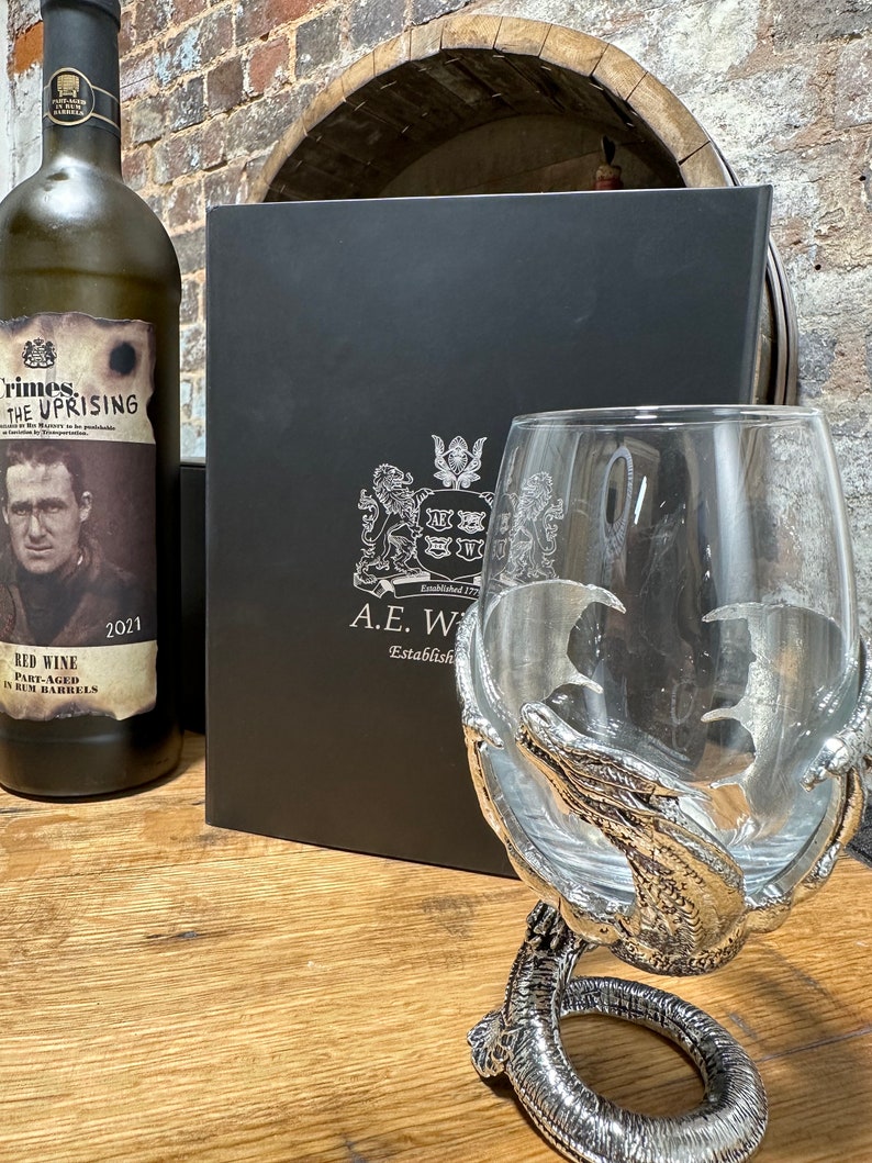May include: A black box with a silver crest and the text "A.E.W. Established 1772" on the front. A silver dragon claw wine glass sits on a wooden surface next to a bottle of red wine with a label that reads "Crimes. The Uprising. Red Wine Part-Aged in Rum Barrels 2021".