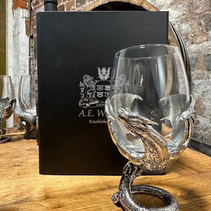 May include: A silver dragon goblet with a clear glass bowl. The goblet is on a wooden surface with a black box and a bottle of wine in the background. The box has the text "A.E.W. Established" on it.