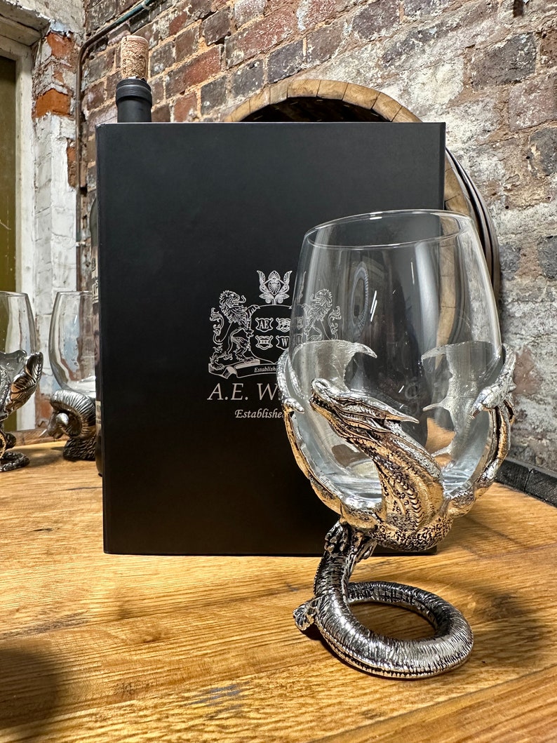 May include: A silver dragon goblet with a clear glass bowl. The goblet is on a wooden surface with a black box and a bottle of wine in the background. The box has the text "A.E.W. Established" on it.