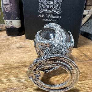 May include: A silver dragon figurine holding a clear glass orb. The dragon is curled around the orb, with its wings spread out. The figurine is sitting on a wooden surface. The text "A.E. Williams Established 1779" is visible on a black box in the background.
