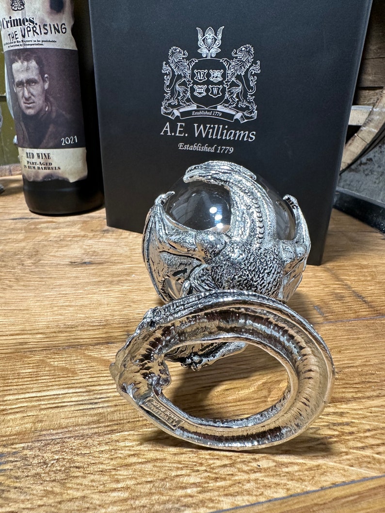 May include: A silver dragon figurine holding a clear glass orb. The dragon is curled around the orb, with its wings spread out. The figurine is sitting on a wooden surface. The text "A.E. Williams Established 1779" is visible on a black box in the background.