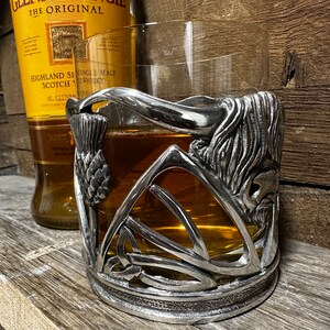 May include: A silver metal whisky glass holder with a Celtic knot design and a thistle. The holder is surrounding a glass of amber colored whisky.