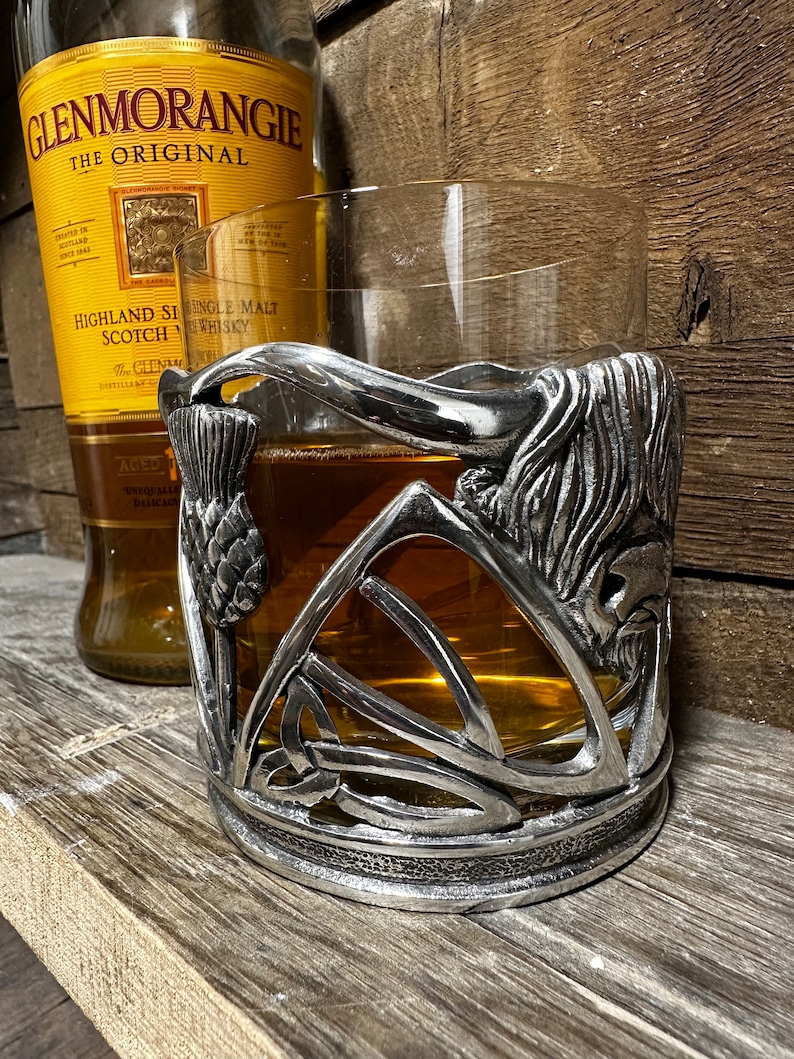 May include: A silver metal whisky glass holder with a Celtic knot design and a thistle. The holder is surrounding a glass of amber colored whisky.