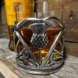 May include: A silver metal drink coaster with a Celtic knot design and a raised thistle in the center. The coaster is designed to hold a glass of whiskey or other beverage.