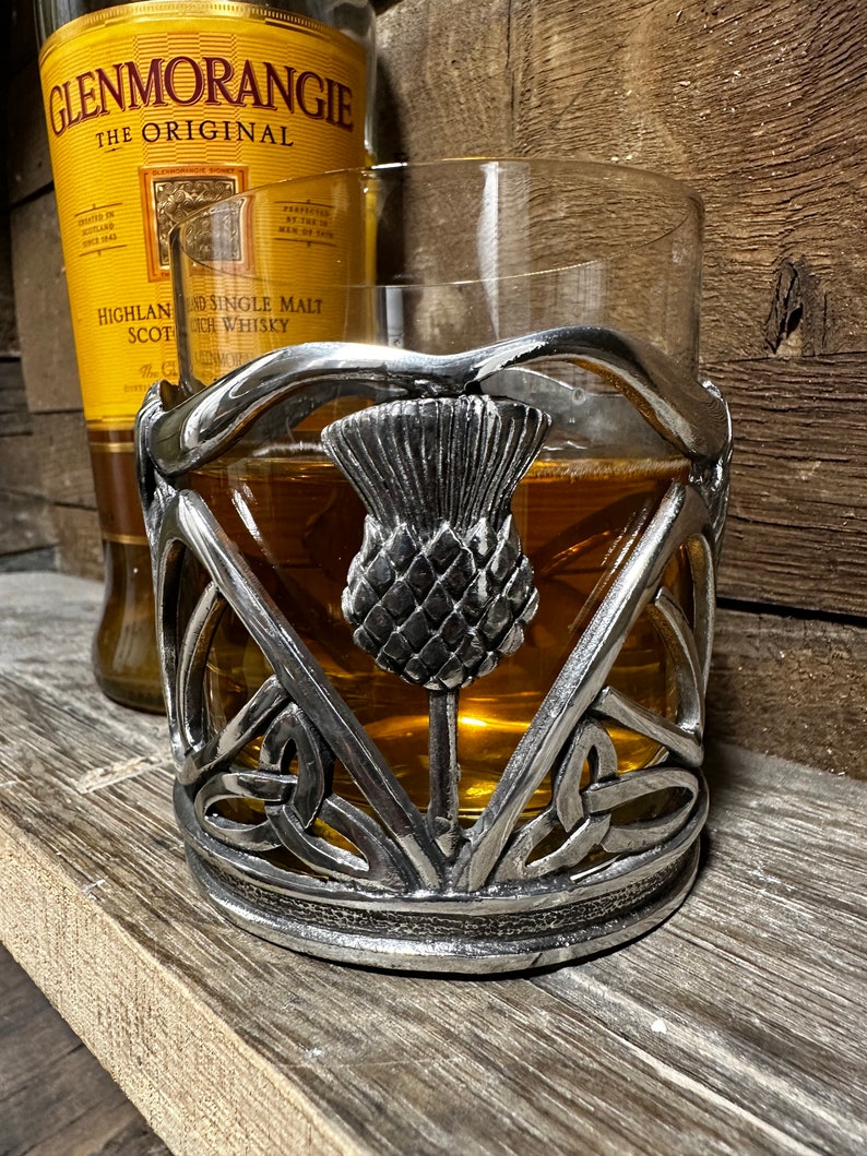 May include: A silver metal drink coaster with a Celtic knot design and a raised thistle in the center. The coaster is designed to hold a glass of whiskey or other beverage.