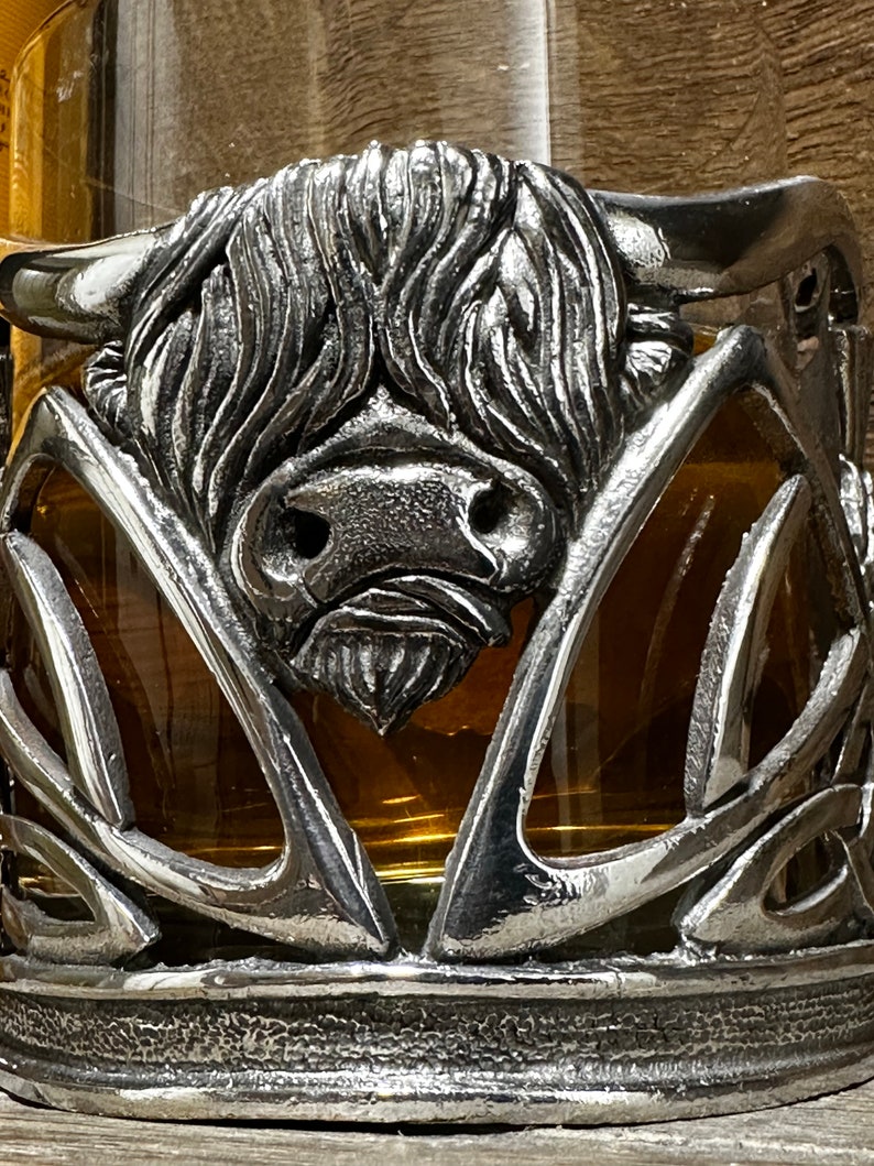 May include: A silver Highland cow head sculpture with intricate details, mounted on a glass cup holder with a Celtic knot design.