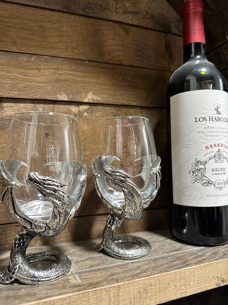 May include: Two silver wine glass holders shaped like dragons, each with a clear glass goblet. The holders are detailed with scales and claws. A bottle of Los Haroldos Reserve Malbec wine is in the background.