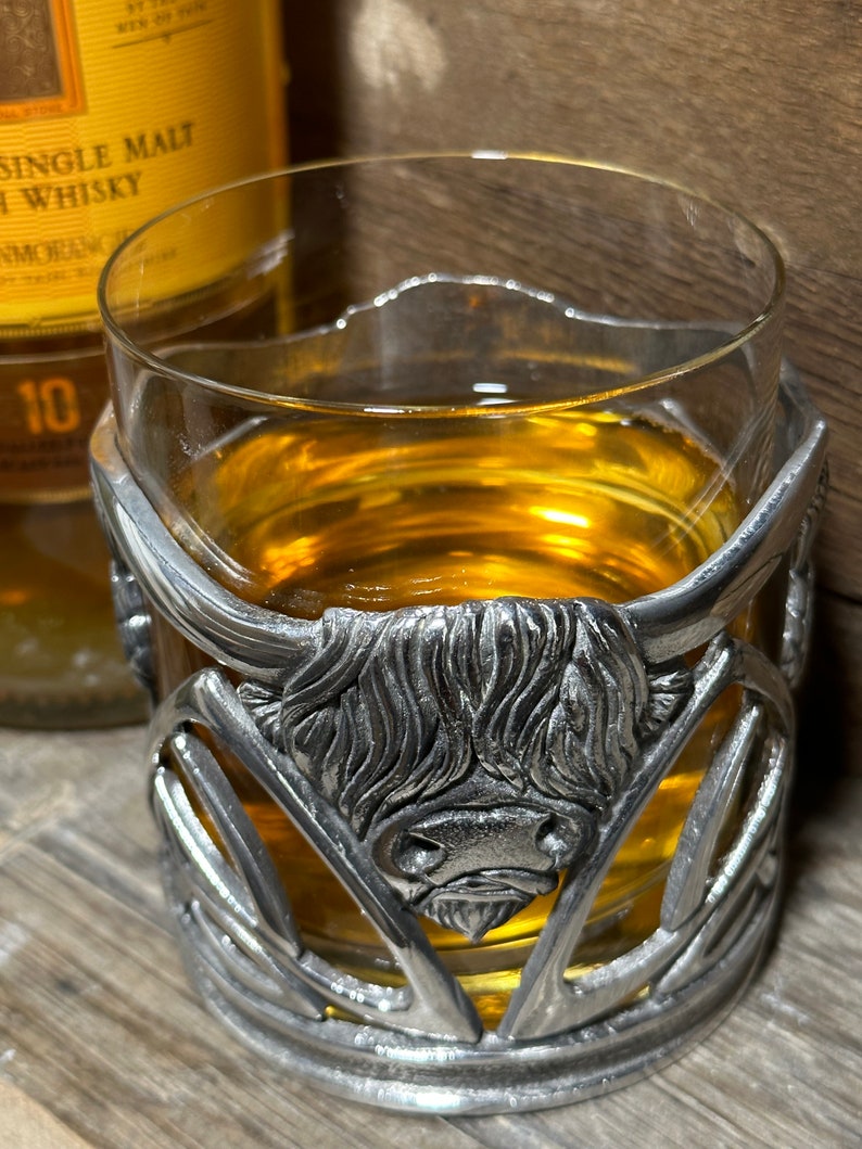 May include: A glass of amber-colored liquid in a clear glass with a pewter Highland cow design coaster. The coaster features a detailed design of a Highland cow's head with horns and a textured fur pattern.
