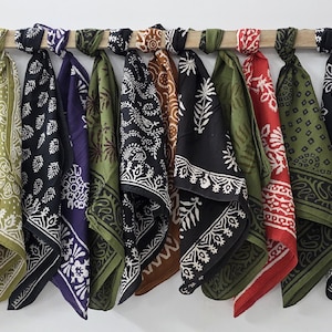 May include: A collection of bandanas in various colors and patterns hanging from a wooden rod. The bandanas feature designs in white on backgrounds of olive green, black, brown, red, and blue. The bandanas are knotted at the top.