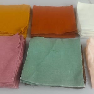 May include: A collection of folded fabric squares in various colors, including yellow, orange, white, pink, green, and peach. The squares are stacked and arranged on a light-colored surface, showcasing a range of hues and textures.