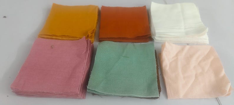 May include: A collection of folded fabric squares in various colors, including yellow, orange, white, pink, green, and peach. The squares are stacked and arranged on a light-colored surface, showcasing a range of hues and textures.