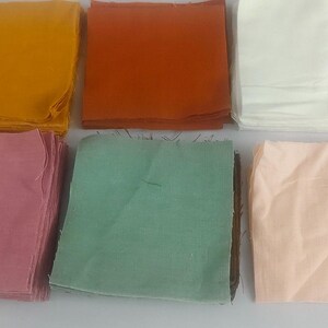 May include: Six folded fabric squares in various colors: mustard yellow, rust orange, white, dusty rose, mint green, and peach. The squares are stacked neatly, suggesting a textile or craft supply.