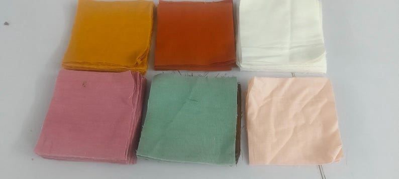 May include: Six folded fabric squares in various colors: mustard yellow, rust orange, white, dusty rose, mint green, and peach. The squares are stacked neatly, suggesting a textile or craft supply.