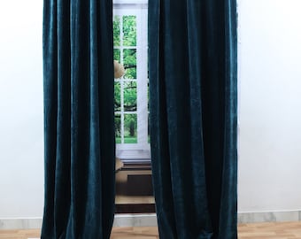 Puede incluir: Dos cortinas de terciopelo verde azulado cuelgan de una barra plateada, enmarcando una ventana con vistas a la vegetación. Las cortinas son largas, con pliegues suaves y un brillo lujoso. La habitación tiene un suelo de madera clara.