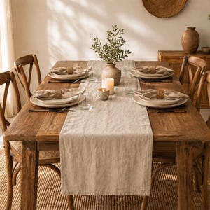 Puede incluir: Una mesa de comedor rectangular de madera preparada para cuatro personas con un camino de mesa de lino beige, platos de cerámica blanca, servilletas de tela beige con anillos trenzados, cristalería transparente, un jarrón y dos velas.