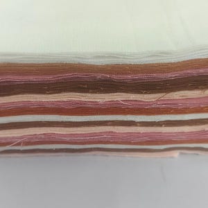 May include: A stack of fabric squares in various shades of white, brown, and pink. The layers are neatly arranged, showcasing a range of colors from light to dark. The fabric appears to be cotton or a similar material, suitable for crafting or sewing projects.