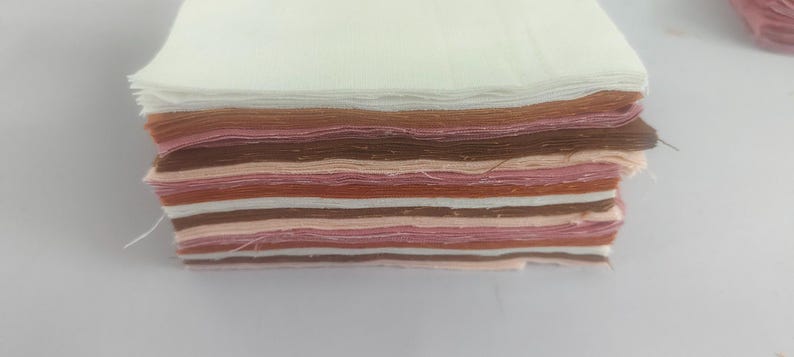 May include: A stack of fabric squares in various shades of white, brown, and pink. The layers are neatly arranged, showcasing a range of colors from light to dark. The fabric appears to be cotton or a similar material, suitable for crafting or sewing projects.