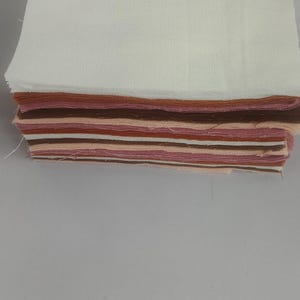 May include: A stack of fabric squares in various shades of brown, pink, and white. The top layer is a large white square, with the other layers showing a mix of colors. Ideal for quilting or crafting.