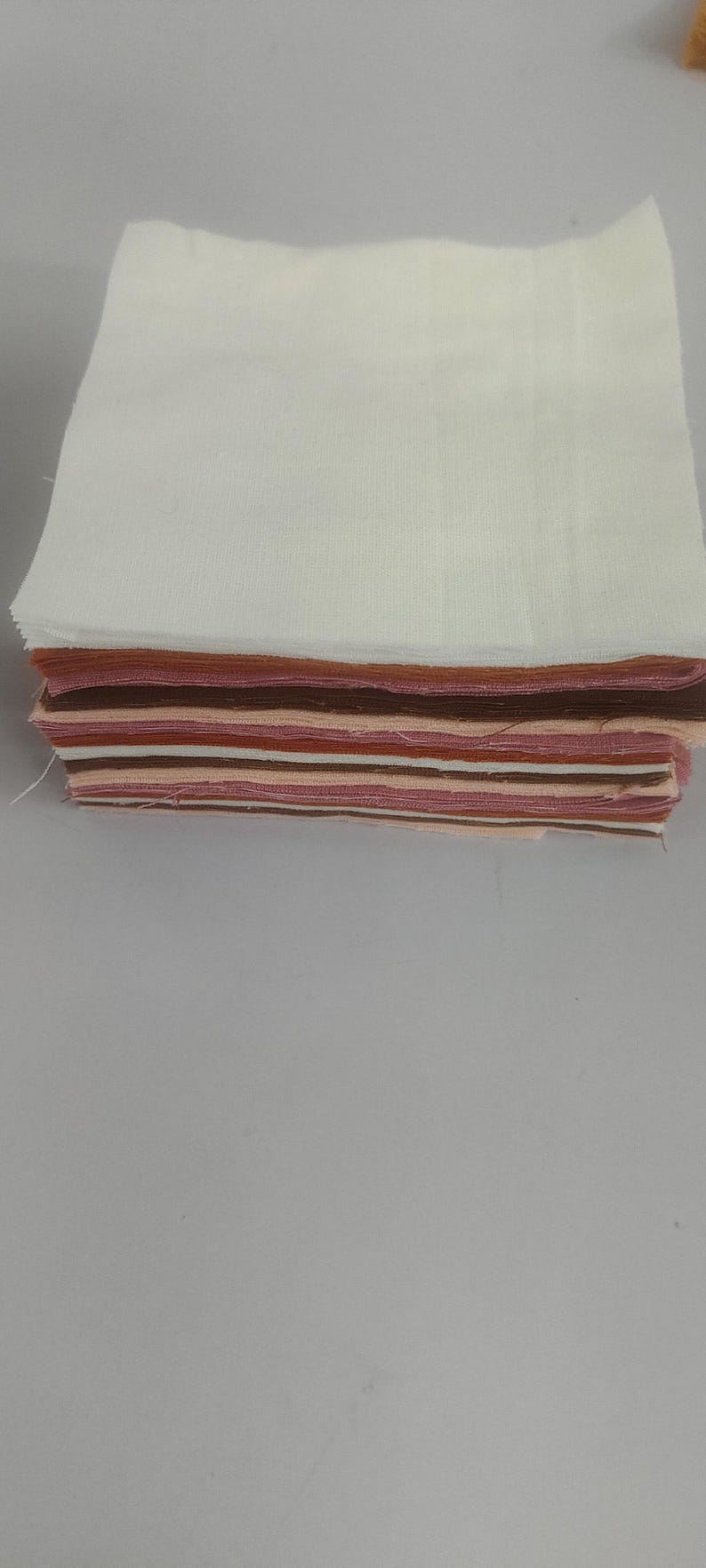 May include: A stack of fabric squares in various shades of brown, pink, and white. The top layer is a large white square, with the other layers showing a mix of colors. Ideal for quilting or crafting.