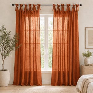 May include: A pair of burnt orange curtains made of a semi-sheer, textured fabric. The curtains are hung on a black rod using fabric ties at the top, creating a gathered look. The panels hang straight down, covering the window frame completely.