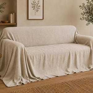May include: A beige sofa cover draped over a couch, featuring a textured fabric that extends over the arms and down to the floor. The cover has a neutral, light tan color and a soft, woven appearance that conforms to the shape of the furniture.