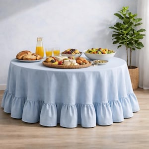 May include: A round, pale blue tablecloth with a frilled edge, set for a meal. The table is laden with food, including bread, cheese, fruit, and salad. A glass bottle of orange juice and two glasses are also on the table. A potted plant is in the background.