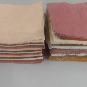 May include: Two stacks of fabric squares in various colors. One stack features a light peach color on top, with stripes of brown, pink, and cream underneath. The other stack has a pink top layer, with layers of white, orange, and yellow visible.