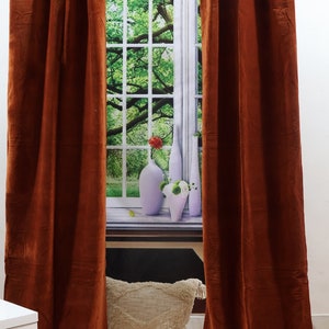 May include: A pair of rust-coloured velvet curtains hang in front of a window with a nature scene. The curtains are fully drawn and the fabric has a soft, luxurious look.