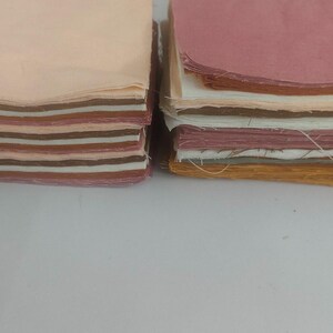 May include: Two stacks of fabric squares in various colors. One stack features shades of peach, brown, and cream. The other stack displays pink, white, and gold tones. The fabric appears to be cotton or a similar material, with visible edges.