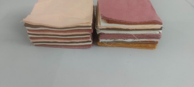 May include: Two stacks of fabric squares in various colors. One stack features shades of peach, brown, and cream. The other stack displays pink, white, and gold tones. The fabric appears to be cotton or a similar material, with visible edges.