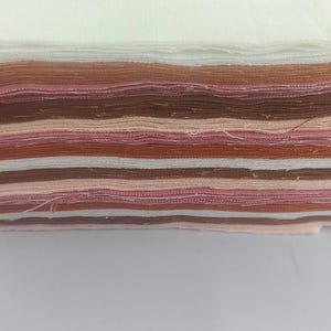 May include: A stack of fabric swatches in various shades of brown, pink, and white. The swatches are layered, showcasing a range of colors and textures. The edges of the fabric are slightly frayed, indicating a handmade or artisanal quality. The fabric appears to be cotton or a similar material.