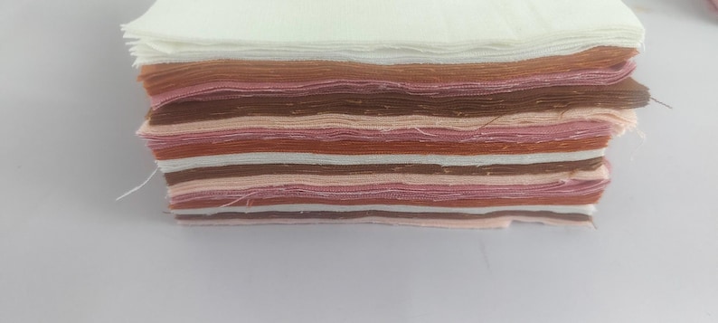May include: A stack of fabric swatches in various shades of brown, pink, and white. The swatches are layered, showcasing a range of colors and textures. The edges of the fabric are slightly frayed, indicating a handmade or artisanal quality. The fabric appears to be cotton or a similar material.